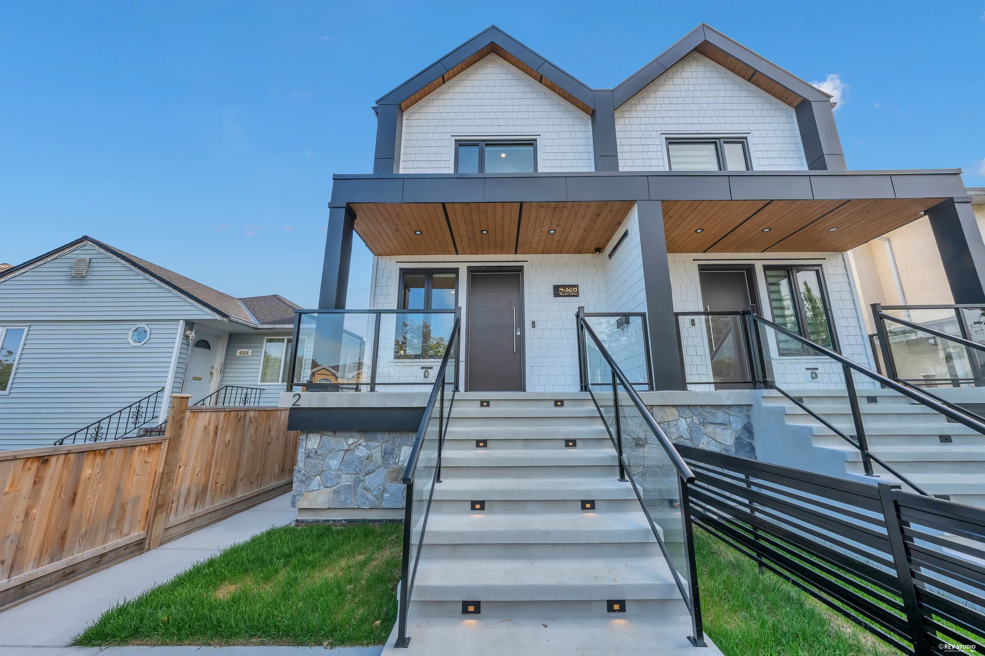 The duplex from the street in daylight: two matching halves, twin gables, cedar-lined porch ceilings over a stone base, black railings on the stairs up, new turf either side