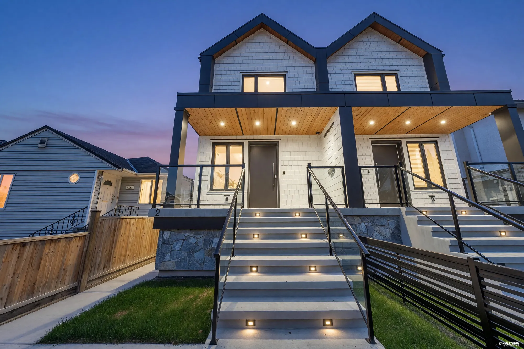 4328 Rupert Street at dusk: a new side-by-side duplex in white board with black trim, cedar-lined porch ceilings over a stone base, both halves lit