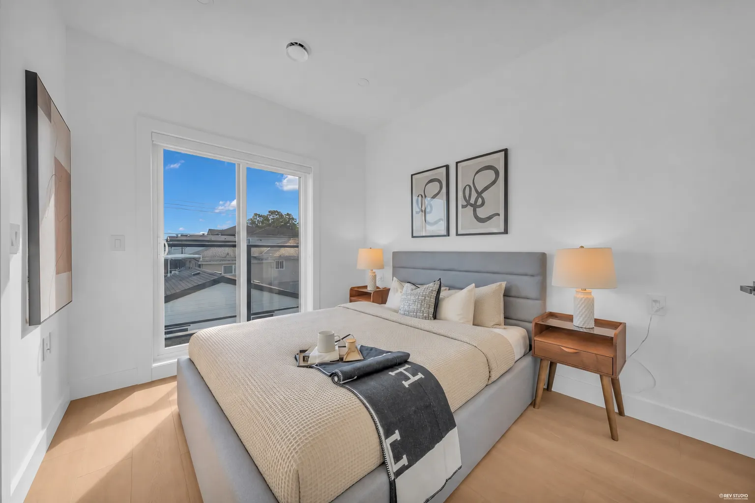 The primary bedroom: a bed with a grey headboard, two framed prints, a lamp on the night table, the doors to the balcony