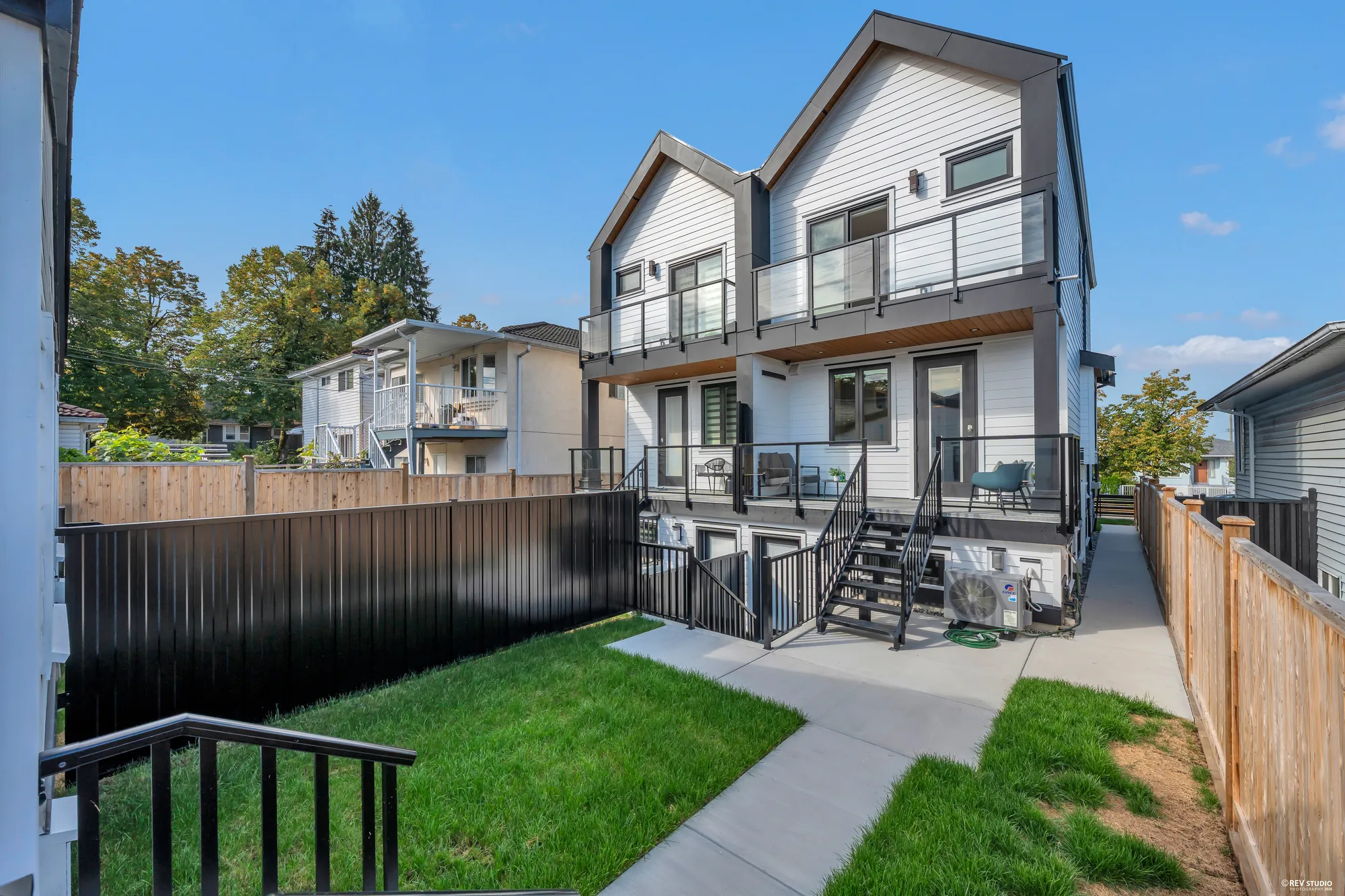 The back of the duplex in daylight: balconies above, covered porches with stairs down, the yards divided by a fence, turf and paths